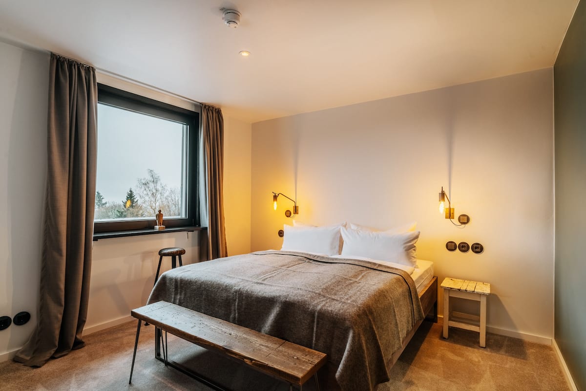 A bedroom with a large bed, a bench, a small table, and a window with curtains.