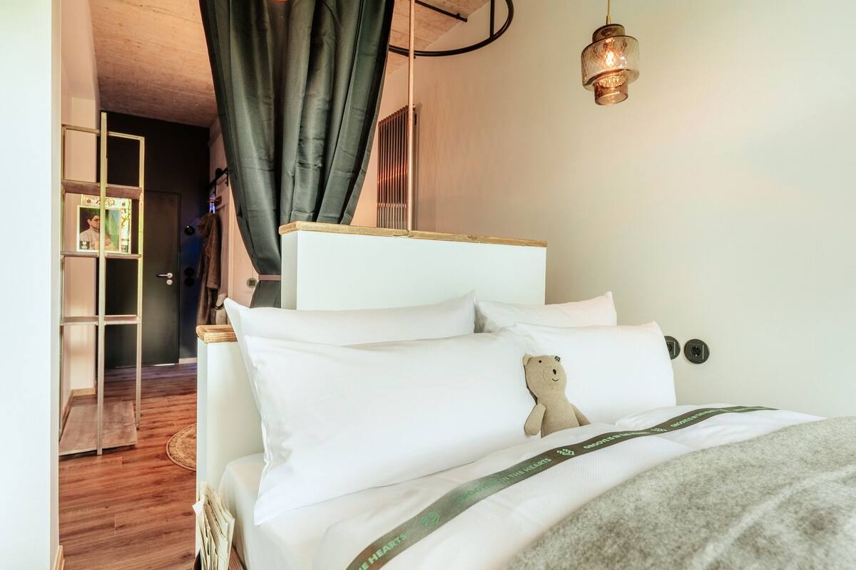 A neatly made bed with white linens and a teddy bear pillow, a hanging light fixture, and a wooden floor.