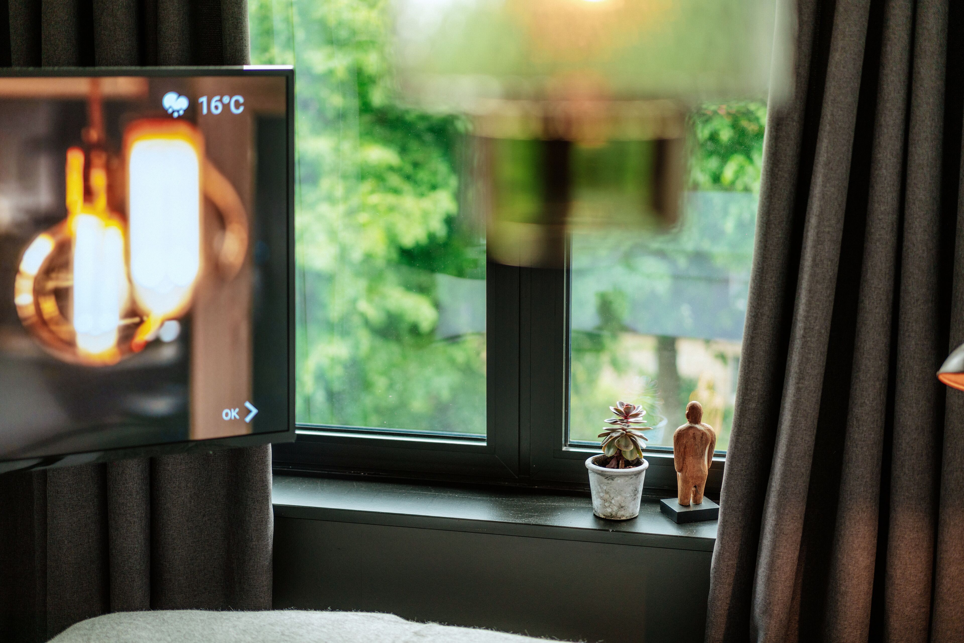 Un televisor que muestra una temperatura de 16°C, una ventana con cortinas, una planta pequeña en maceta y una figurita en el alféizar.