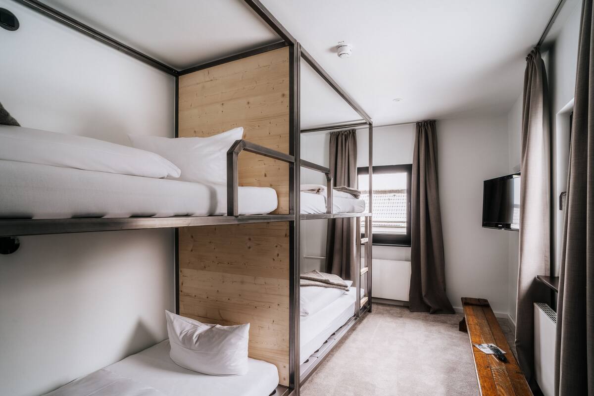 A bunk bed room with a wooden panel wall, a bench, a TV, and a window with curtains.