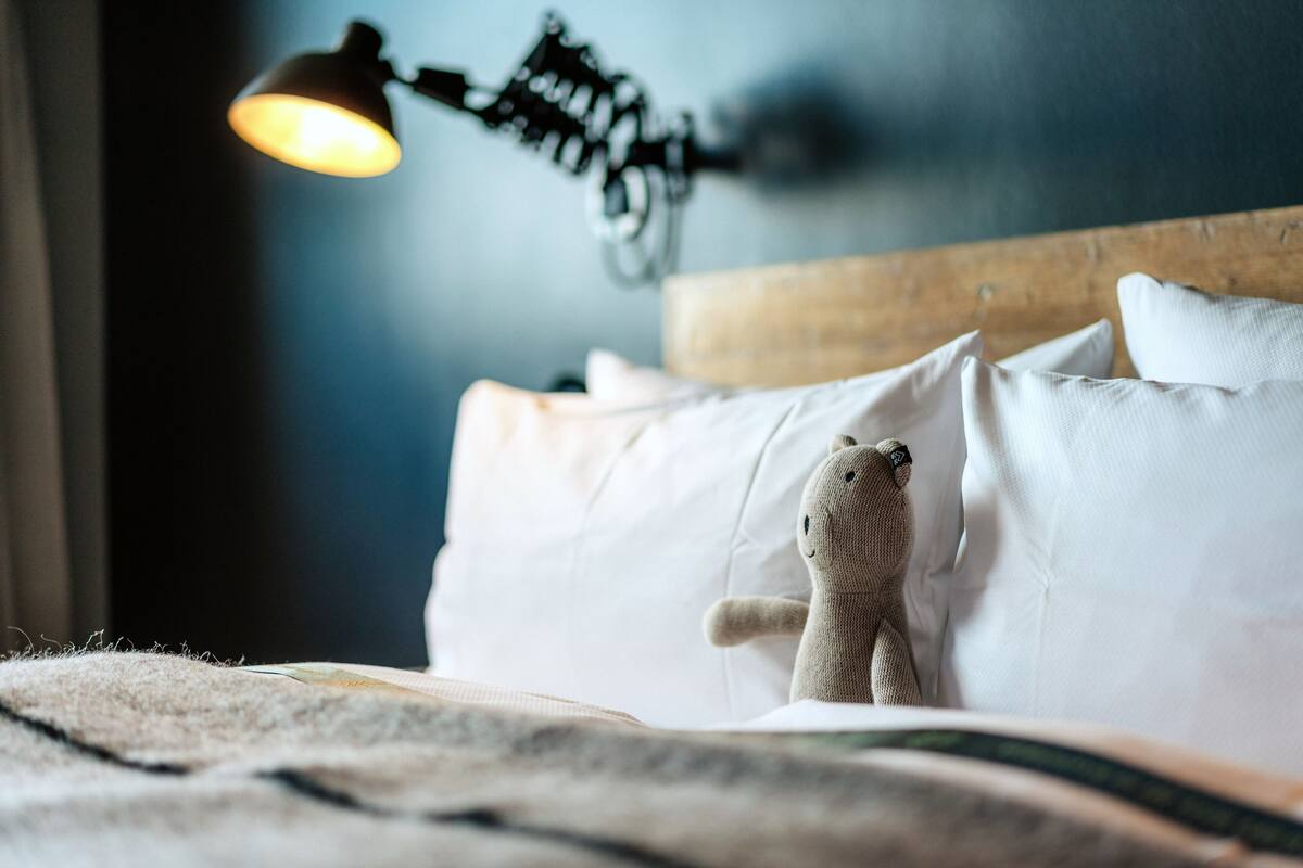 A bed with white bedding, a teddy bear, and a wall-mounted lamp.
