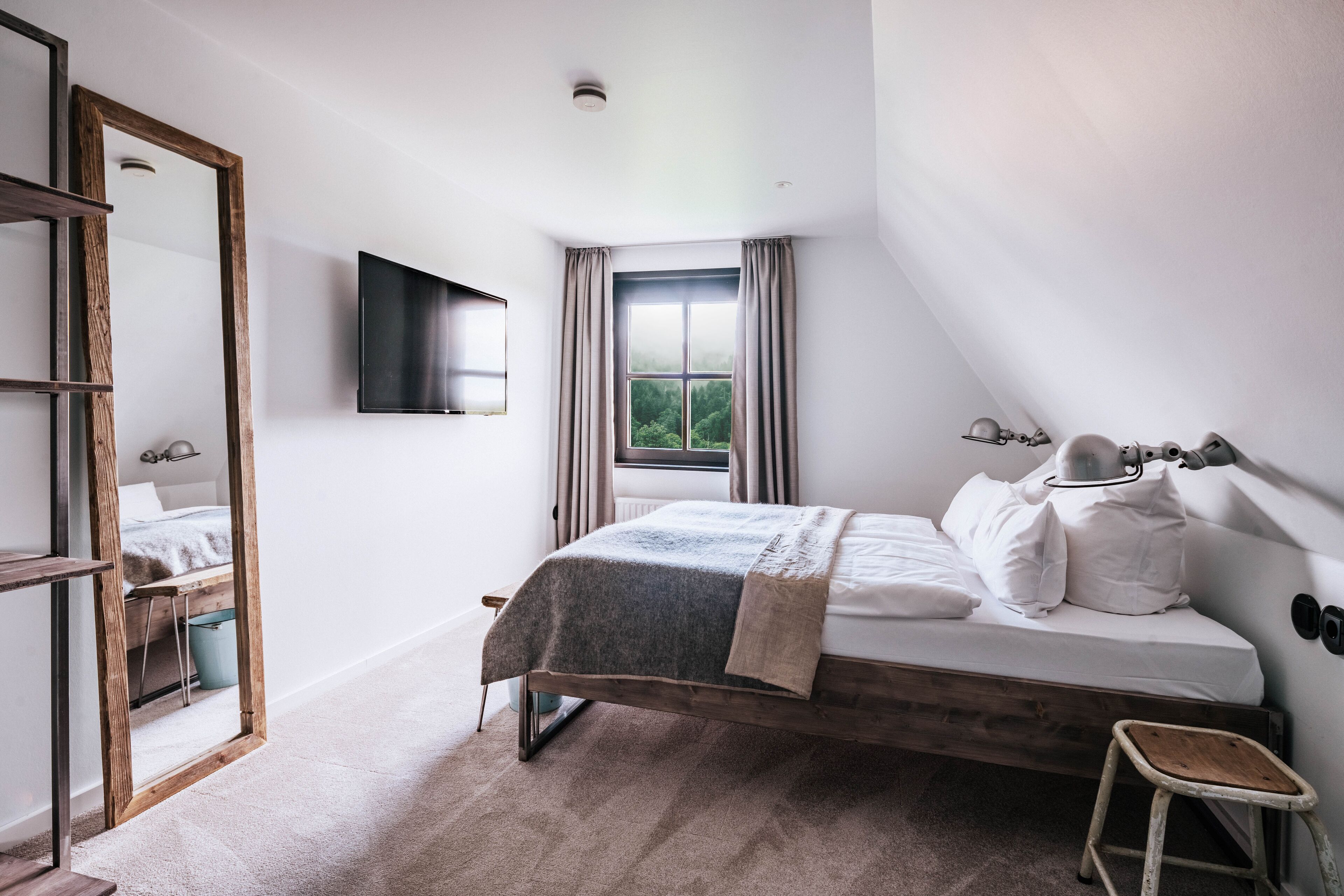A bedroom with a bed, a TV mounted on the wall, a window with curtains, and a mirror.