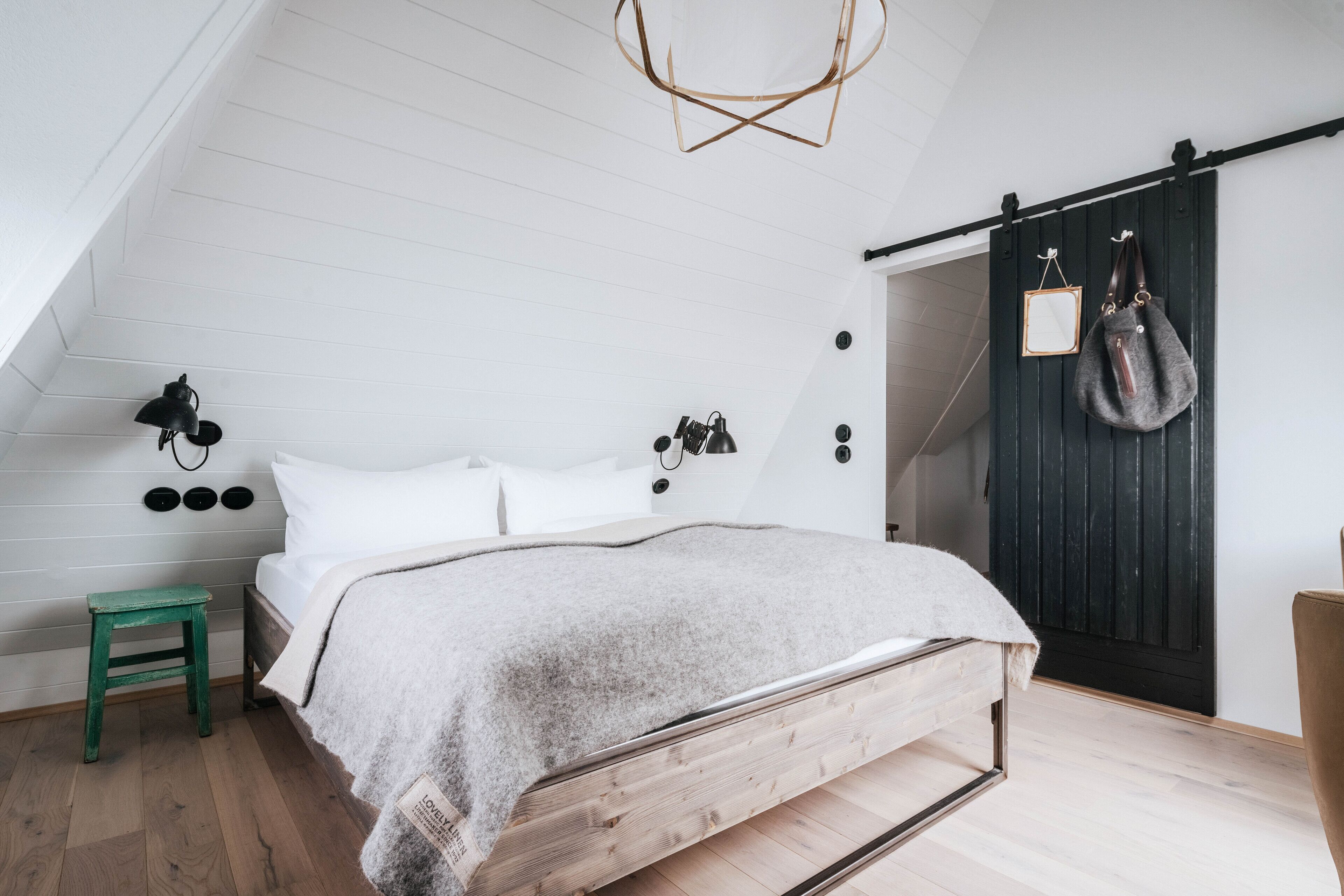 Een slaapkamer met een schuine balkenlaag, een bed met houten frame, een groene kruk, een wandlamp en een donker houten deur met spiegel en een tas eraan.