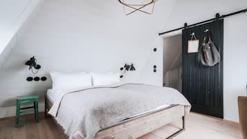 Een slaapkamer met een schuine balkenlaag, een bed met houten frame, een groene kruk, een wandlamp en een donker houten deur met spiegel en een tas eraan.