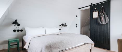 A bedroom with a sloped ceiling, a bed with a wooden frame, a green stool, a wall-mounted lamp, and a dark wooden door with a mirror and a bag hanging on it.