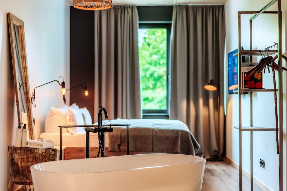 A modern bedroom with a large bathtub, a bed with white linens, a wooden nightstand, and a window with curtains.