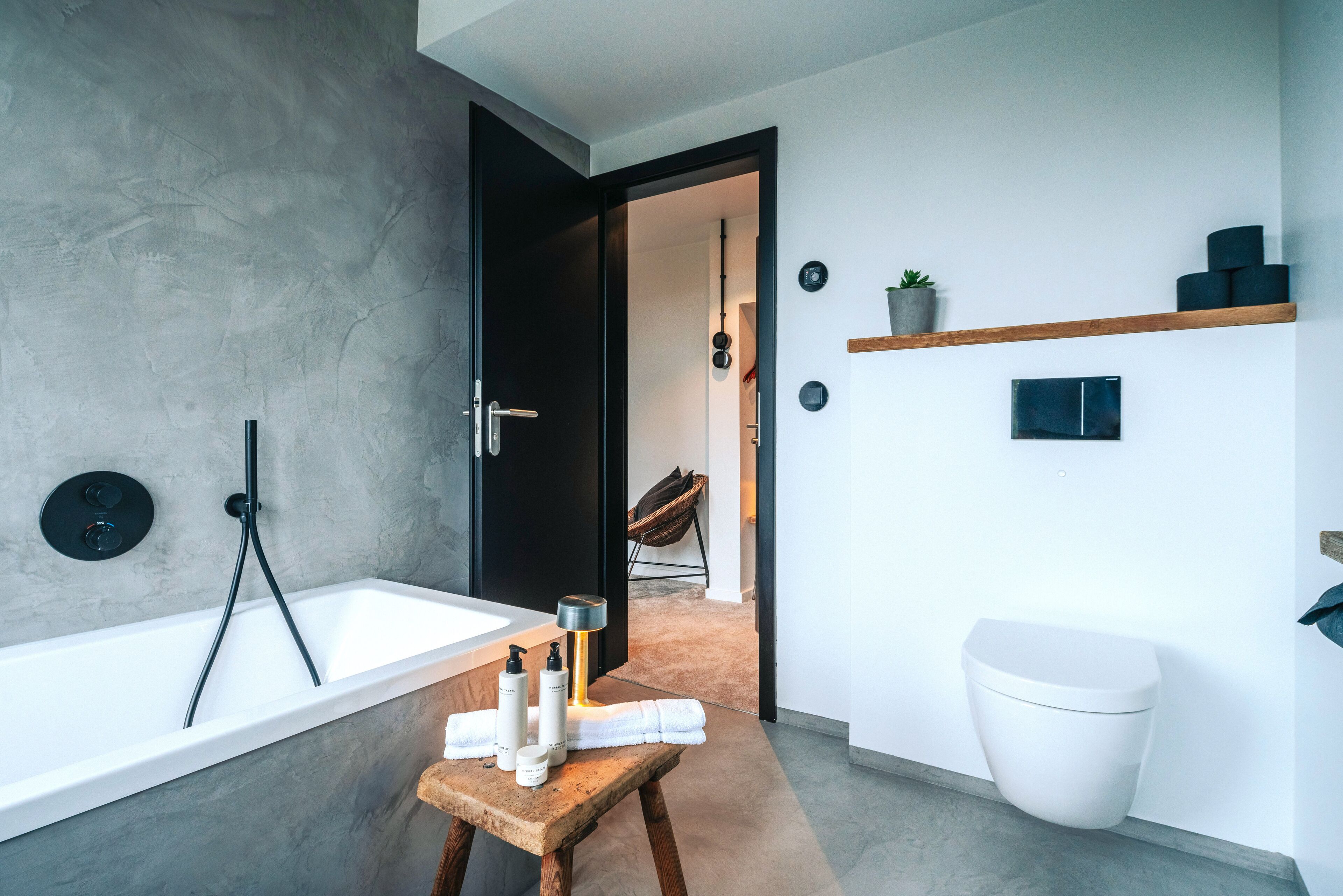 Ein modernes Badezimmer mit einer Badewanne, einem hölzernen Hocker mit Pflegeprodukten, einer Toilette und einem Regal mit einer Pflanze.