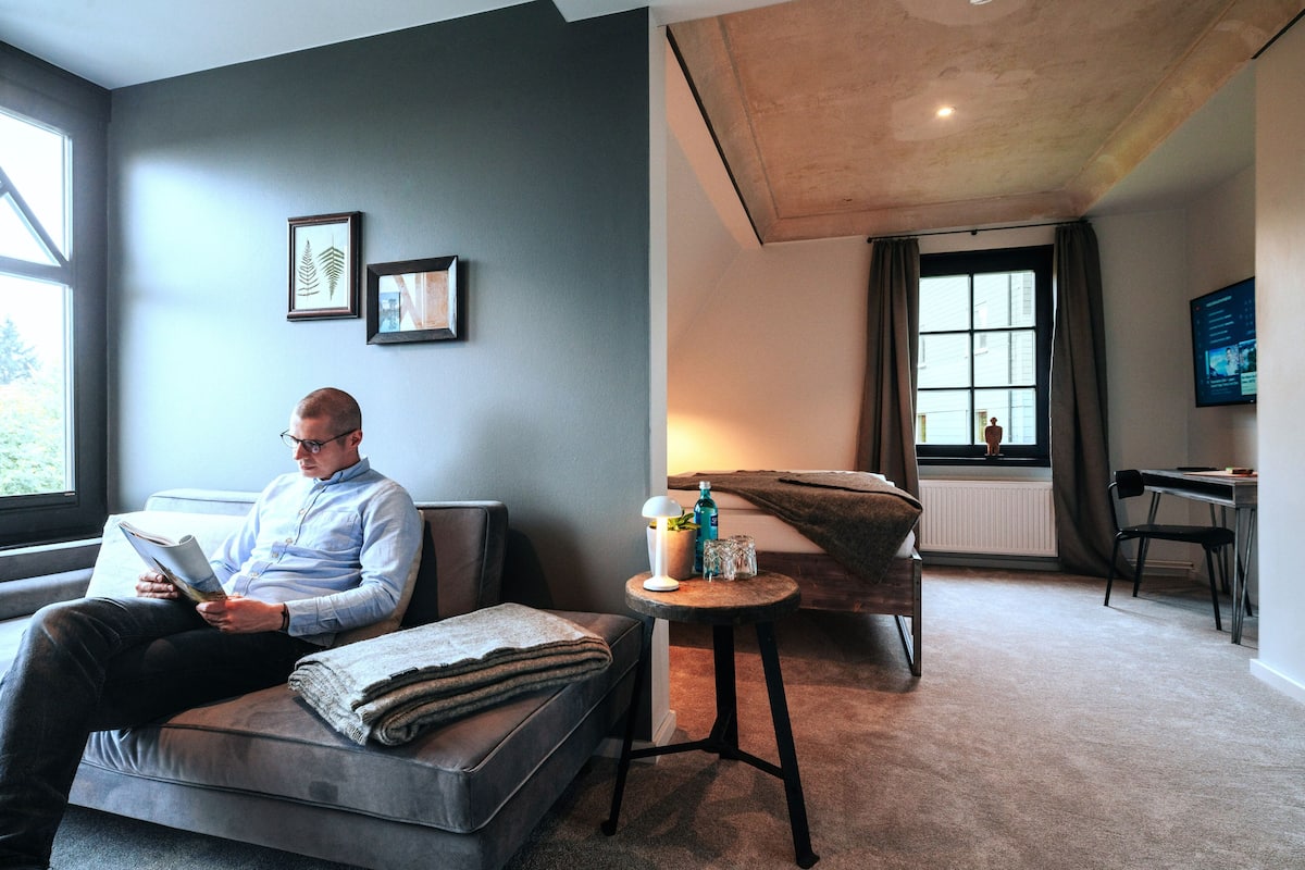 A modern hotel room with a large window, a sofa, a small table with a lamp, and a bed with a blanket.