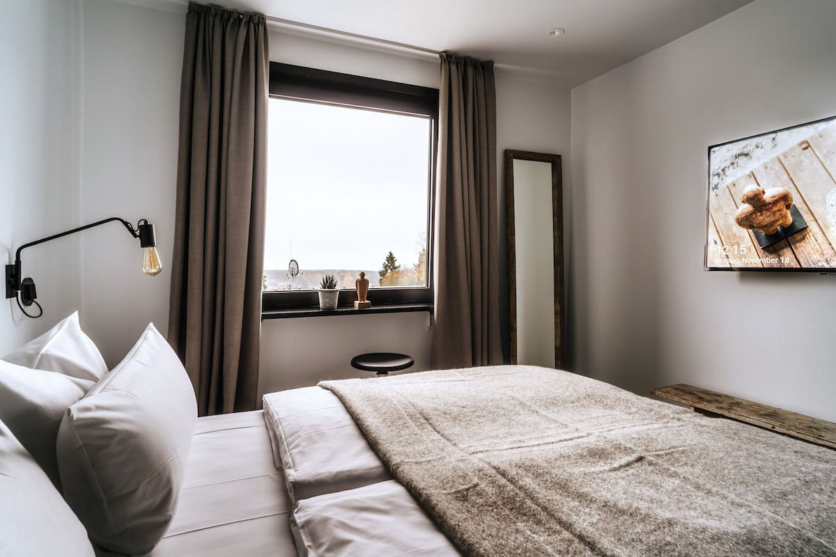 A modern hotel room with a large bed, a bedside lamp, a window with curtains, a small table with a plant, and a wall-mounted TV displaying a date and time.