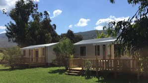 A mobile home with a wooden deck and a small porch, surrounded by greenery and a mountain in the background. - Camping Milella (Olmeto)