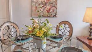 Een eetgedeelte met een ronde tafel, vier stoelen, een kroonluchter, een aan de muur hangend schilderij en een lamp.