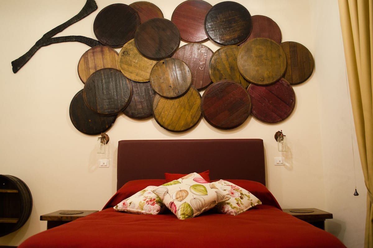 Uma cama com colcha vermelha e almofadas florais, parede com mosaico circular de madeira, e cabeceira de madeira escura.