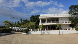 A beachfront hotel with a two-story structure, a covered outdoor seating area, and palm trees.