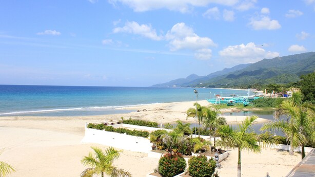 A beach with a view of the ocean and mountains, a white building with a garden, and boats in the water.