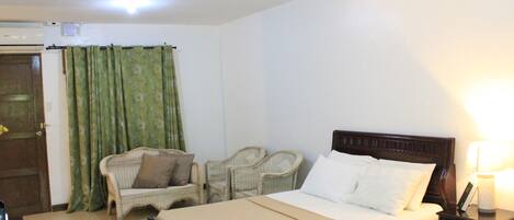 A hotel room with a bed, two chairs, a bedside table with a lamp, and a door with curtains.