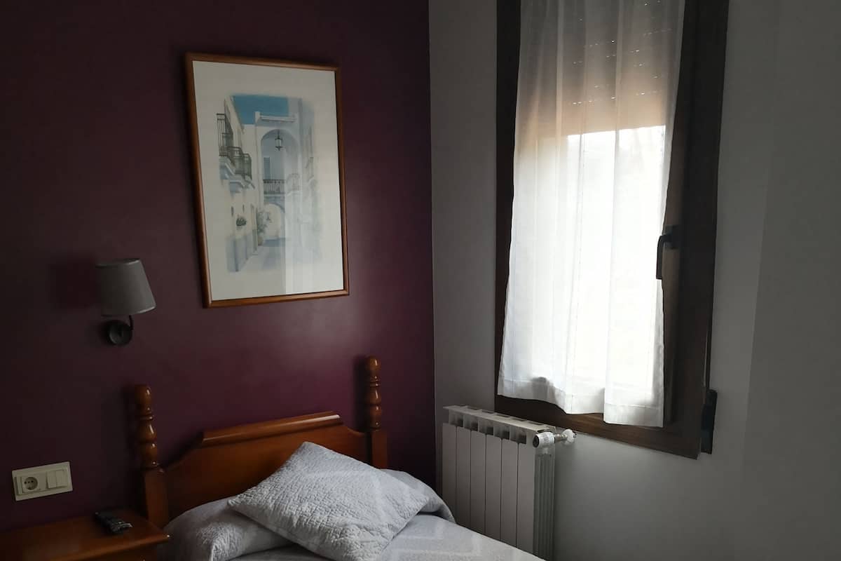 Un dormitorio con una cama individual, una mesita de noche, una lámpara de pared, un cuadro enmarcado y una ventana con cortinas translúcidas.