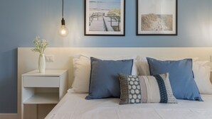 Un dormitorio ordenado con cama, almohadas, una mesita de noche, un florero y dos cuadros en la pared.