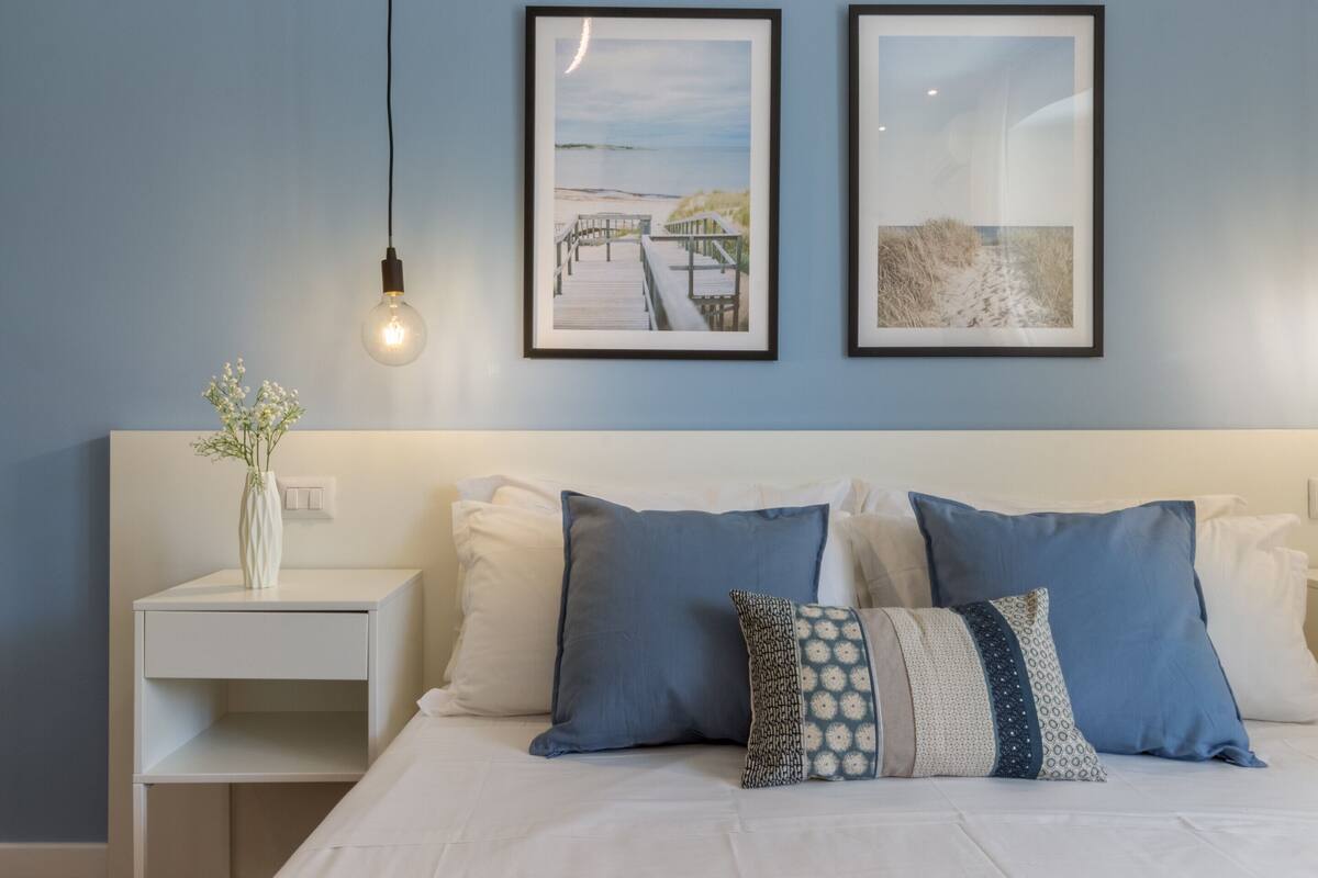 A neatly arranged bedroom with a bed, pillows, a nightstand, a vase, and two framed pictures on the wall.