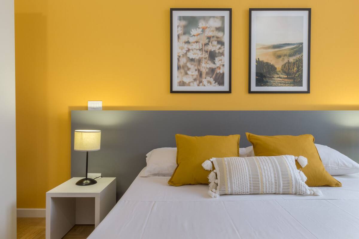 A neatly made bed with white bedding and two mustard pillows, a bedside lamp, and two framed pictures on the wall.