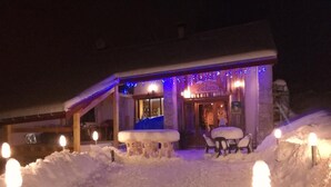 Un espace extérieur aménagé avec des tables et des chaises, recouvert de neige, éclairé par des projecteurs au sol, devant un bâtiment doté d’un toit en pente et décoré de guirlandes lumineuses.