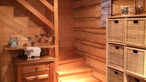 L’intérieur d’un chalet en bois, avec un escalier en bois, une petite armoire en bois dotée de tiroirs, des paniers en osier sur une étagère et une paire de chaussures posées sur le sol.