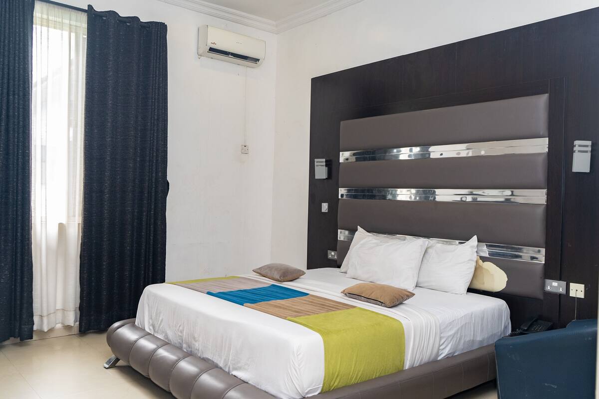 Quarto de hotel moderno com uma cama grande, cabeceira com ripas metálicas, mesa de cabeceira e telefone embutido.