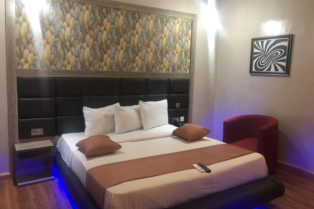 Quarto de hotel com uma cama grande, cabeceira com design texturizado, uma poltrona vermelha e uma obra de arte na parede.