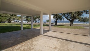 A covered outdoor area with a concrete floor, white pillars, and a view of a grassy park and body of water.