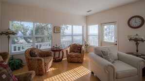 A living room with a sofa, two armchairs, a small table, and a lamp. There are large windows with blinds, a clock on the wall, and a framed picture on the wall.