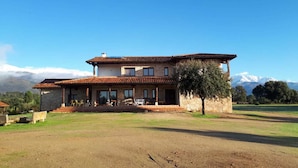 A two-story stone house with a tiled roof, a covered patio with outdoor furniture, and a view of snow-capped mountains in the background. - Sakura Vera (Villanueva De La Vera)