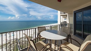 Un balcon avec une table ronde et des chaises, donnant sur une plage parsemée de parasols et offrant une vue dégagée sur l’océan.