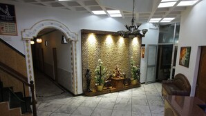Un vestíbulo con piso de mármol, mostrador de recepción, escalera y pared decorativa con estatuas y plantas.