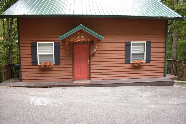 Cozy cabin with a red front door, surrounded by a wooden deck and a paved driveway.