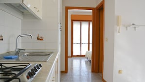 A kitchen with a gas stove, sink, and cabinets. There is a hallway leading to a room with a bed. - Residence Club Azzurro (Massignano)