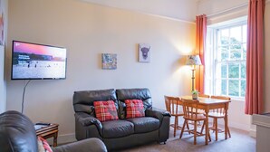 Ein Wohnzimmer mit einer Ledersofa, einem an der Wand montierten Fernseher, einem kleinen Tisch mit Stühlen und einem Fenster mit Vorhängen.