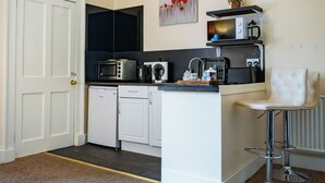 Eine kompakte Küche mit Mikrowelle, Toaster und Kaffeemaschine. Eine weiße Tür, ein kleiner Kühlschrank und ein Barhocker sind vorhanden.