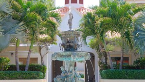 Una fontana con statue in un cortile circondato da palme e un edificio con tetto in tegole rosse.
