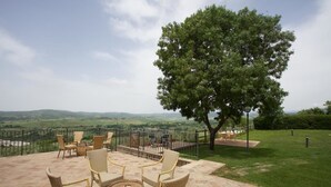 Een terras buiten met rieten meubels en een ronde tafel, met uitzicht op een mooi landschap met heuvels en een grote boom.
