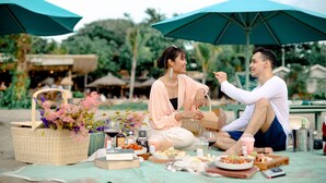 A picnic setup with a wicker basket, food, drinks, and a camera on a table under a large umbrella. - Paisa Seminyak Living (Keroboken Kelod)