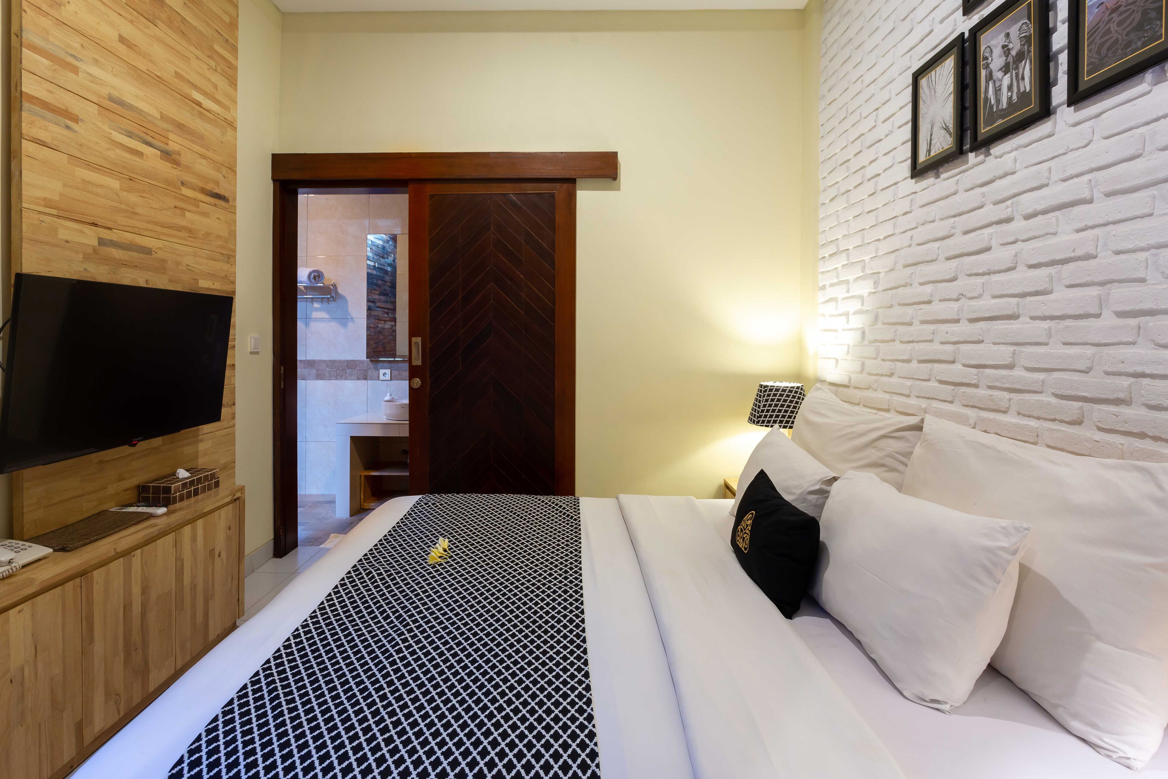 A hotel room with a wooden panel wall, a flat-screen TV, a bed with white bedding and a black and white patterned bedspread, a wooden nightstand, and a white brick wall with framed pictures.