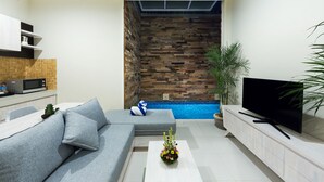 A modern living room with a grey sofa, a flat-screen TV, a white coffee table with flowers, a wooden wall, and a small pool. - Paisa Seminyak Living (Keroboken Kelod)