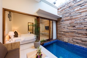 A modern hotel room with a large window, a bed, a bedside table with a lamp, a potted plant, and a view of a swimming pool. - Paisa Seminyak Living (Keroboken Kelod)
