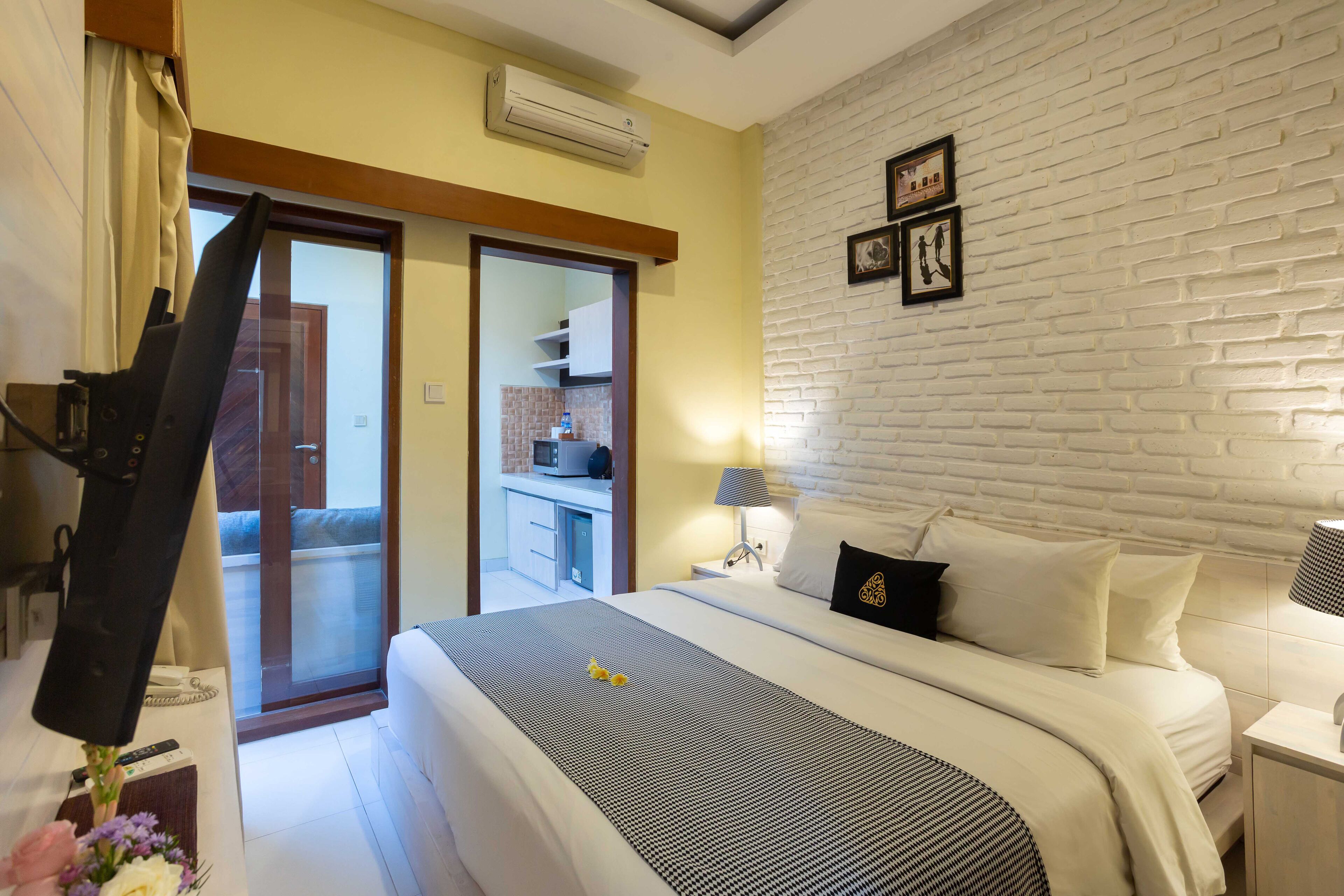 A hotel room with a bed, a desk, a chair, a lamp, and a view of the kitchen and balcony.