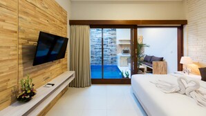 A modern hotel room with a wooden accent wall, a flat-screen TV, a bed with towels, a small seating area, and a view of a pool and outdoor space. - Paisa Seminyak Living (Keroboken Kelod)