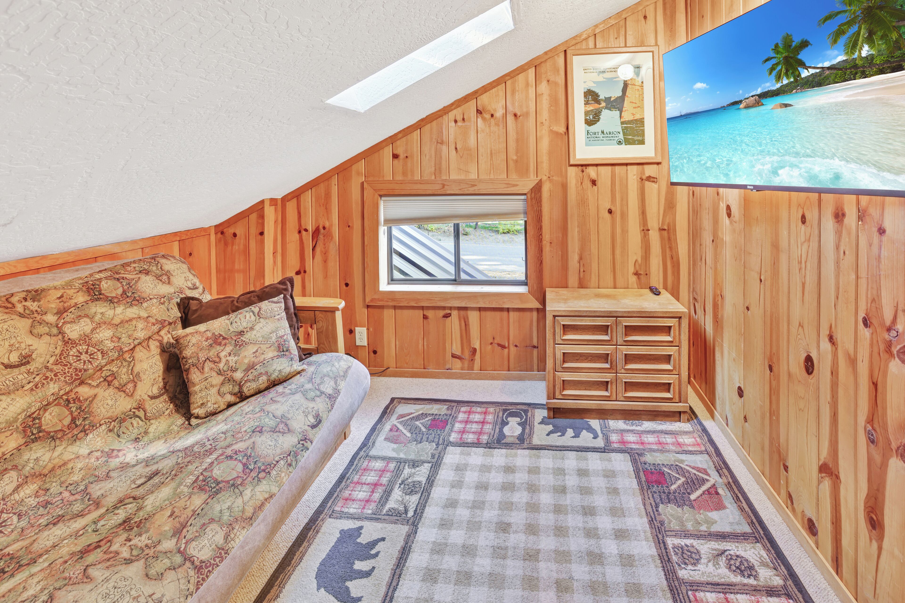 A bedroom with a wooden sloped ceiling, a large window, a bed with patterned bedding, a small wooden cabinet, and a framed picture of a beach scene.