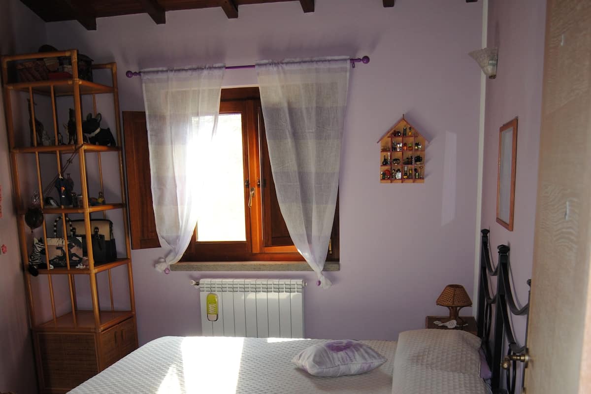 A bedroom with a bed, wooden floor, a window with curtains, a radiator, a shelf with items, and a small table.