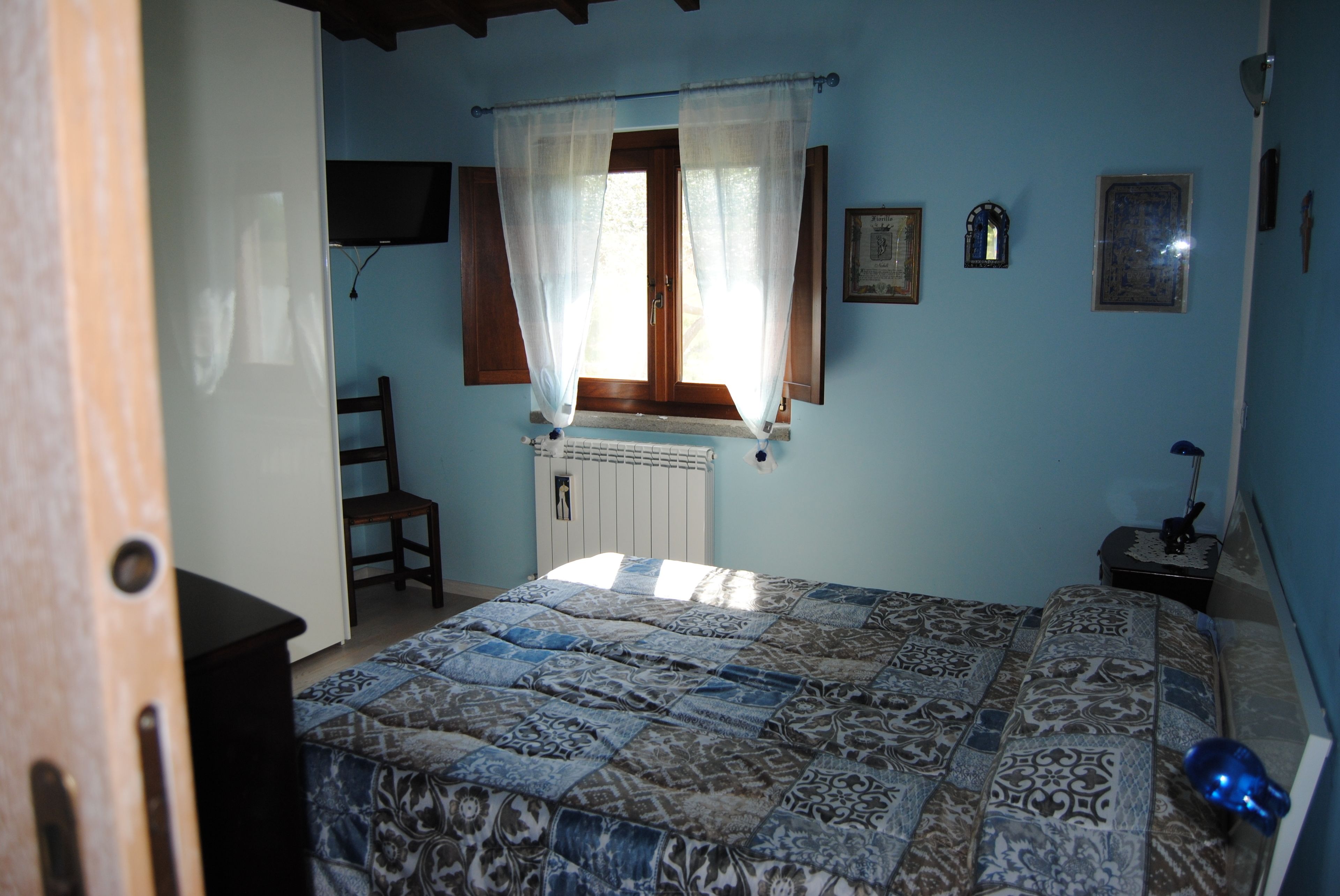 A bedroom with a bed, a window with curtains, a radiator, a chair, a desk, and a lamp.