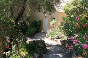 A narrow stone pathway leading to a building with a door, surrounded by lush greenery and flowering plants. - Les Asphodèles (Partinello)