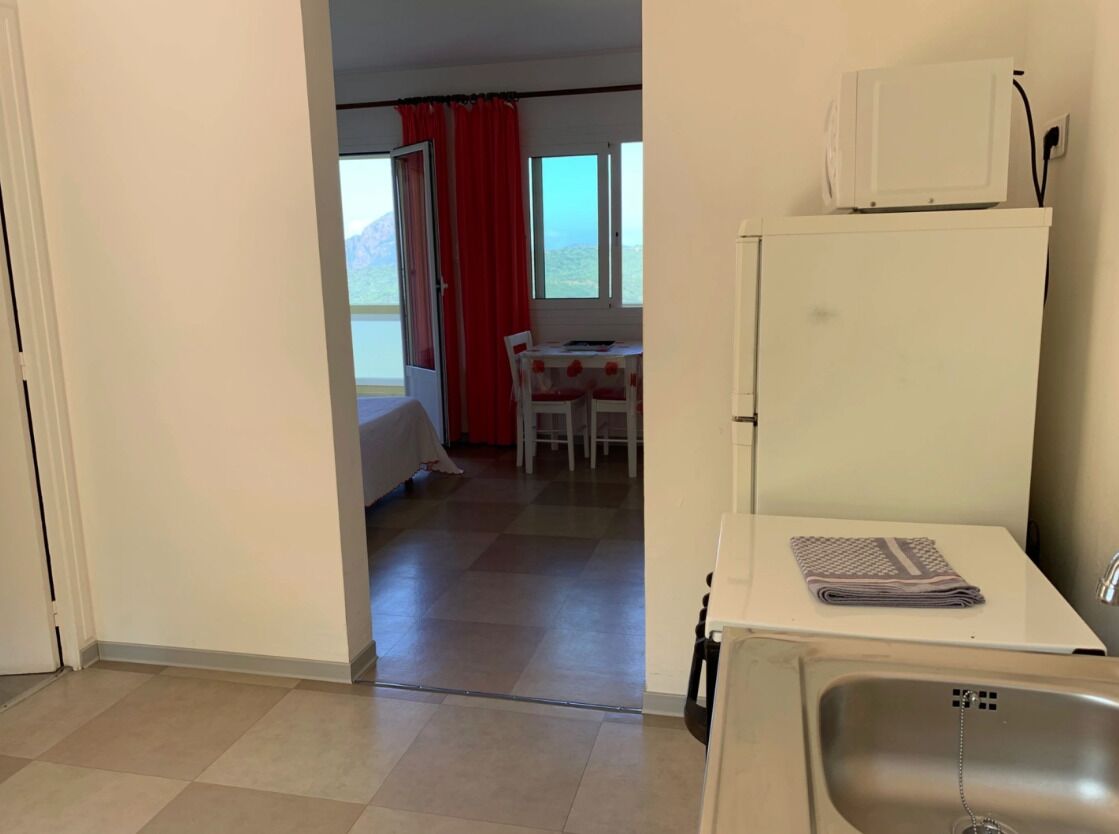 A compact hotel room with a kitchenette, a bed, a dining area with a table and chairs, and a view of the mountains through the window.