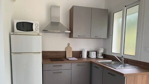 A compact kitchen with a microwave, stove, and sink. It features light wood cabinets and a window with frosted glass. - Les Asphodèles (Partinello)